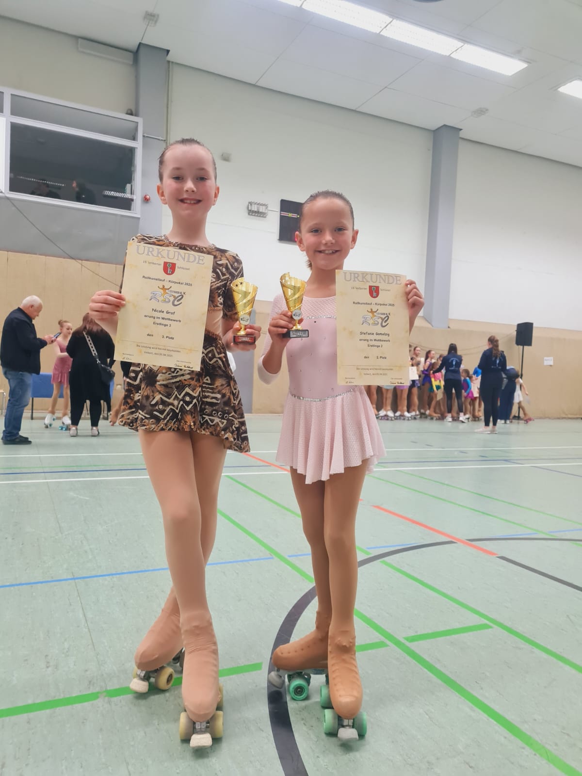 Eine Gruppe von elf jungen Rollkunstläuferinnen in glitzernden Kürkleidern und ein Trainer stehen lächelnd in einer Sporthalle.