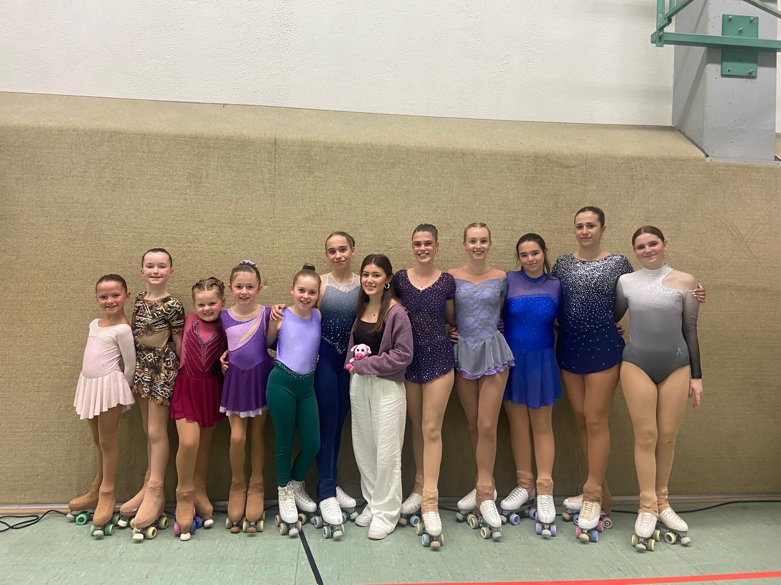 Eine Gruppe von elf jungen Rollkunstläuferinnen in glitzernden Kürkleidern und ein Trainer stehen lächelnd in einer Sporthalle.