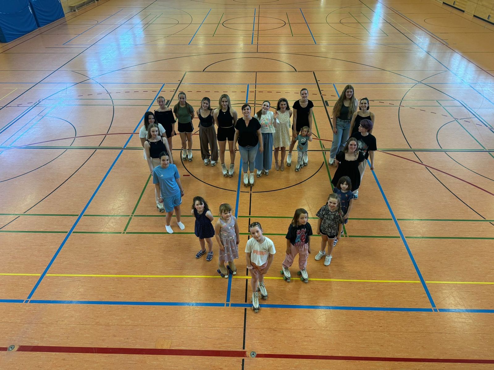 Eine große Gruppe von Rollkunstläuferinnen unterschiedlichen Alters hat sich in einer Sporthalle zu einer Herz-Formation aufgestellt.